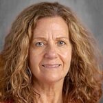 A woman with curly hair and a warm smile, wearing an orange floral top, against a gray background.