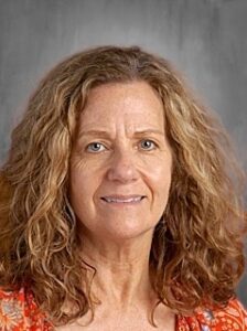 A woman with curly hair and a warm smile, wearing an orange floral top, against a gray background.