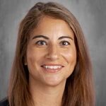 Smiling woman with long brown hair, wearing a black top, against a gray background.