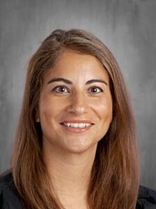 Smiling woman with long brown hair, wearing a black top, against a gray background.