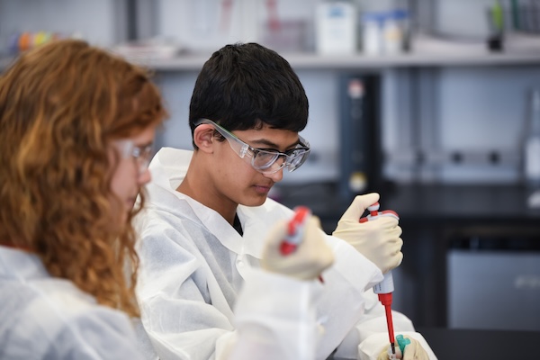 Two students in lab coats and goggles are working in a laboratory, using pipettes for an experiment.