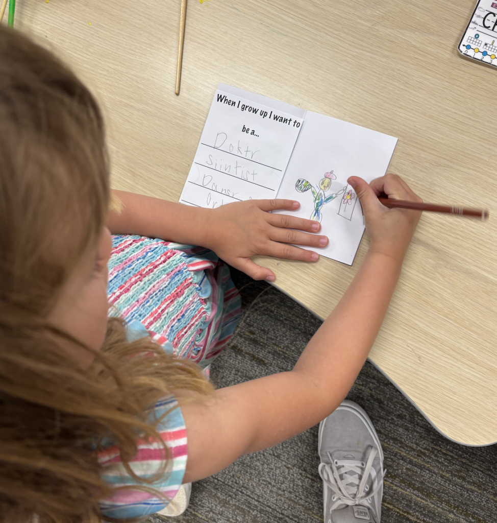 A child writes and colors a drawing in a booklet titled "When I grow up I want to be..." on a tabletop.