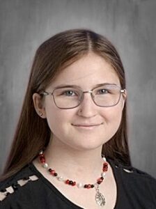 A young person with long brown hair, glasses, and a beaded necklace smiles against a gray background.