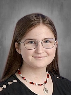 A young person with long brown hair, glasses, and a beaded necklace smiles against a gray background.