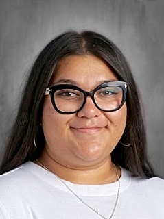 A smiling individual with long dark hair, wearing large black glasses and a white shirt, against a gray background.