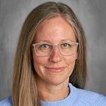 A person with long hair and glasses, wearing a light blue sweater, smiles at the camera against a gray background.
