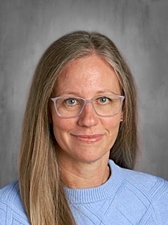 A person with long hair and glasses, wearing a light blue sweater, smiles at the camera against a gray background.