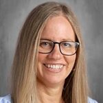 A woman with long blonde hair and glasses smiles warmly, wearing a light blue top against a gray background.