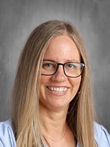 A woman with long blonde hair and glasses smiles warmly, wearing a light blue top against a gray background.