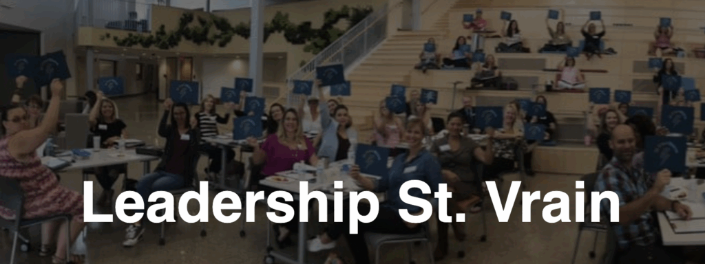 A group of people in a large room raise blue signs, engaged in a Leadership St. Vrain event.
