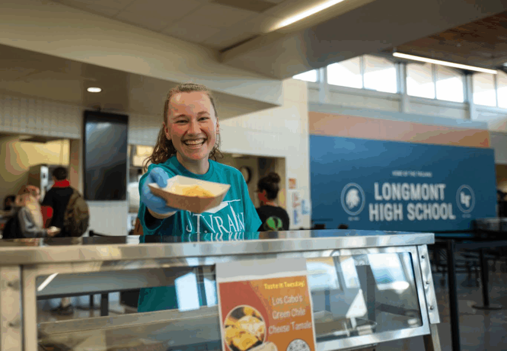 A smiling student serves tamales at Longmont High School's cafeteria, with other students in the background.