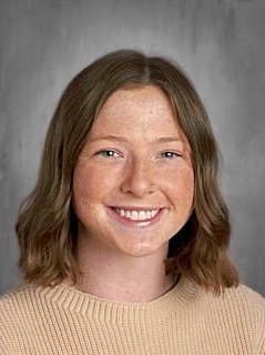 Smiling person with shoulder-length brown hair wearing a beige sweater against a gray background.
