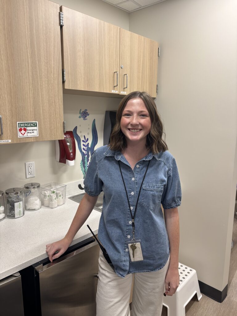 A smiling woman in a denim shirt stands in a kitchen area with cabinets, a sink, and medical supplies visible.