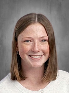 A smiling young woman with straight, shoulder-length brown hair, wearing a light sweater against a gray background.
