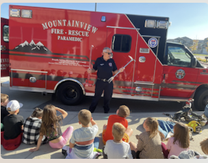 A firefighter speaks to a group of children seated in front of a red fire rescue vehicle.