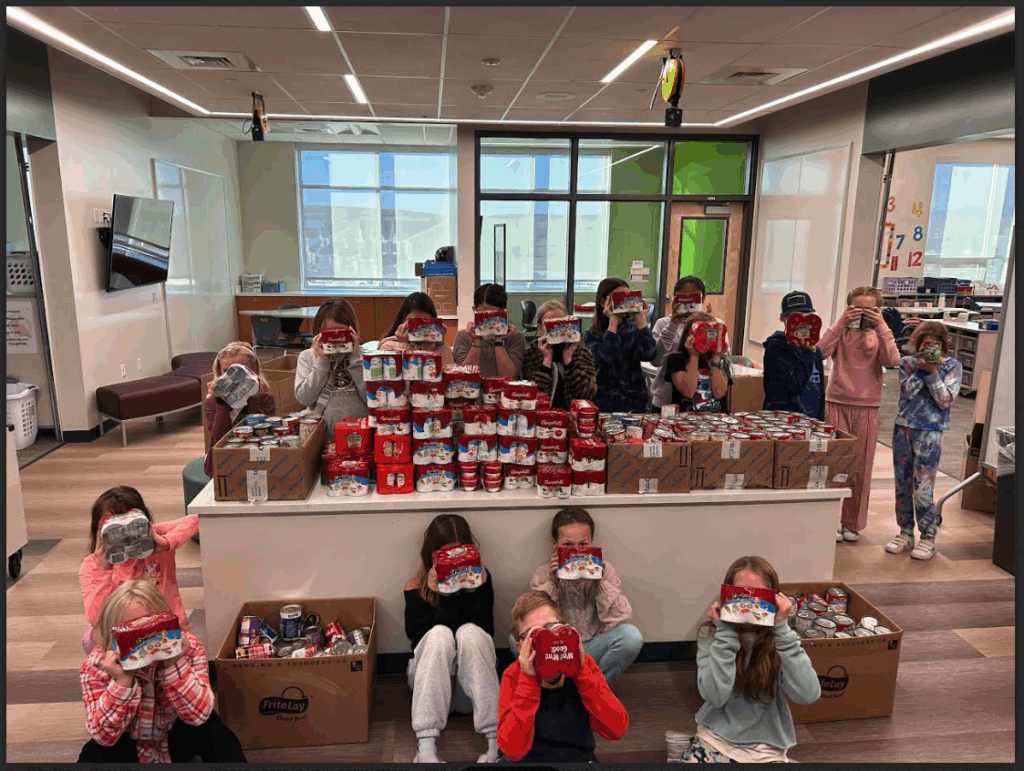 A group of children holds food cans while posing in a classroom filled with stacked boxes of donations.