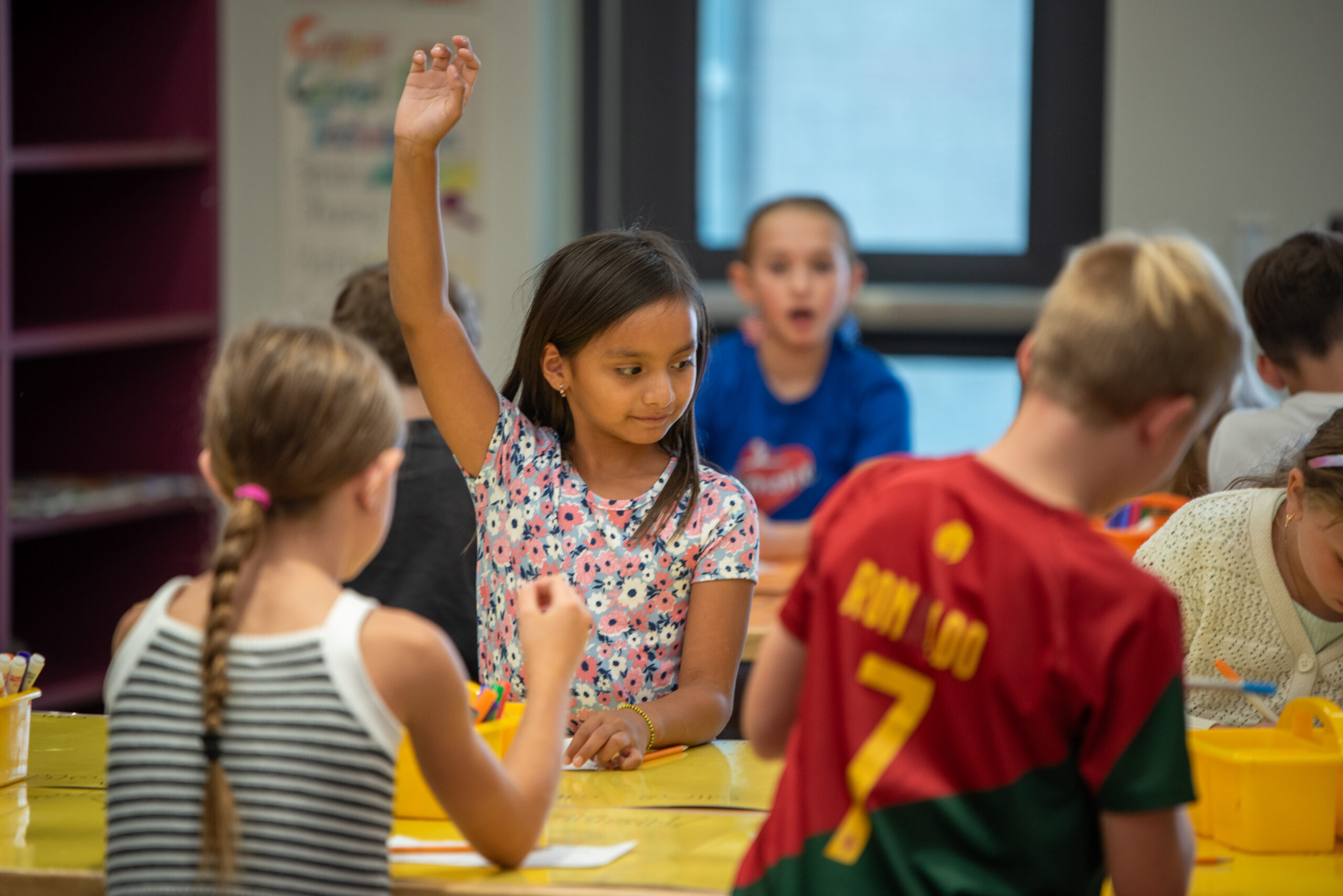 A girl with raised hand participates in a classroom activity, surrounded by classmates engaged in various tasks.