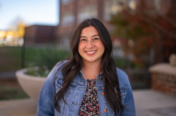 A young woman with long dark hair smiles while wearing a denim jacket over a floral top, outdoors in a garden setting.