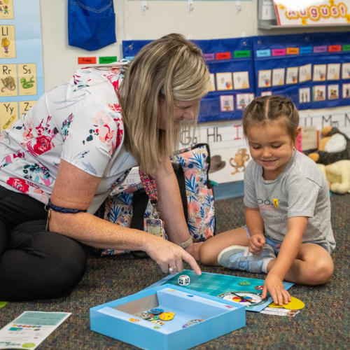 A teacher and a young girl are playing a board game on the floor in a colorful classroom filled with educational materials.