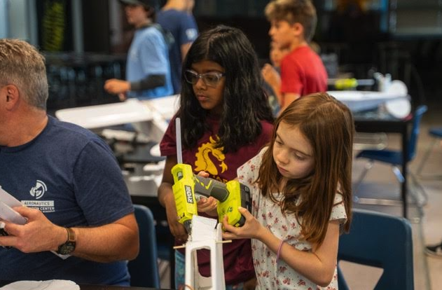 Two children work on a project at a table, using tools while adults assist in a classroom setting.