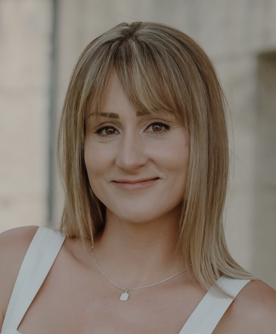 A smiling woman with shoulder-length blonde hair, wearing a white top and a silver necklace, is in focus.