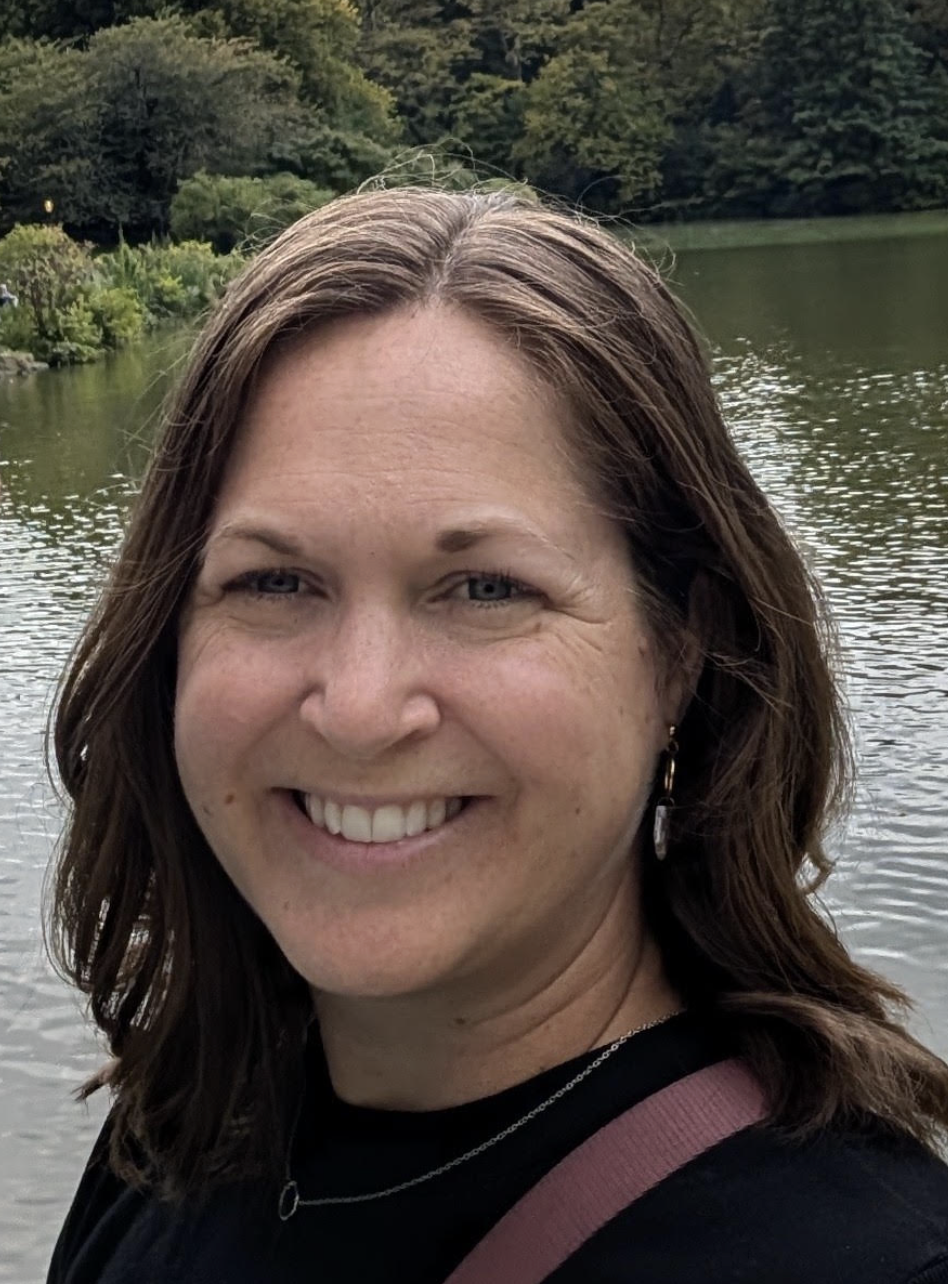 Smiling woman with shoulder-length brown hair stands by a lake, surrounded by greenery and trees in the background.